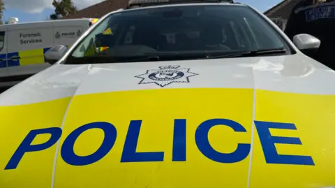 A close up of the bonnet of a police car. The wording "police" is on the bonnet of a car in the foreground. A marked police forensic services van is in the background. 