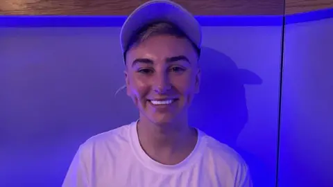 Joshua wearing a white t-shirt and white baseball cap. He has white teeth and blonde hair with dark eyebrows. He is stood in front of a wall which is lit in blue light.