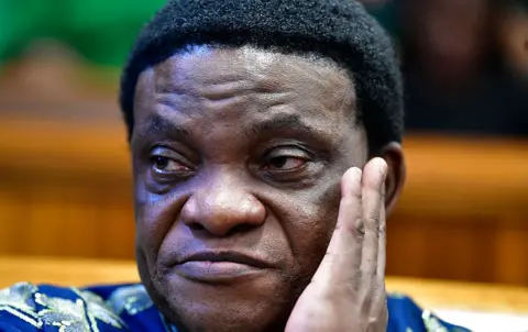 Gallo Images via Getty Images A close-up photo of Timothy Omotoso holding his head in one hand, wearing a blue patterned shirt