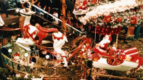 Trevor Wilson A large indoor Christmas display suspended above a shopping centre atrium. At the centre is a decorated Christmas tree and hanging beside it is a festive display featuring a model of Santa Claus seated in a red sleigh filled with presents. In front of the sleigh, a large model reindeer is suspended in mid‑air as if pulling it.