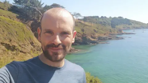 Sam Burns Sam Burns wears a blue T-shirt and has balding, dark hair and a dark goatee beard. He stands in front of a lake with hills to the side of it with trees dotting them
