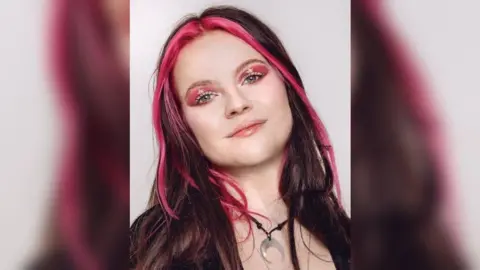 A formal mid-shot of Millie who is smiling to camera and has long black hair with two pink streaks down either side of her face, and bright pink eye makeup