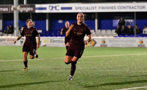 Ipswich Town FC Sophie Peskett wearing a burgundy kit and celebrating while holding up three fingers 