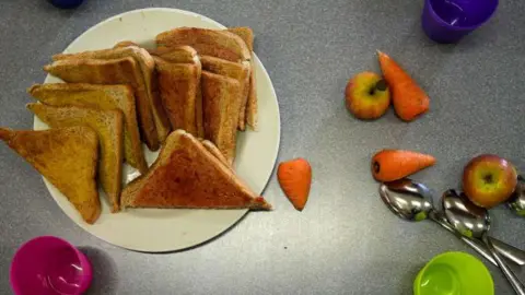 Getty Images A white plate with slices of buttered toast sitting on it. Next to the plates are three multi-coloured glasses of water. There are mini carrots, two apples and silver spoons also on the table.
