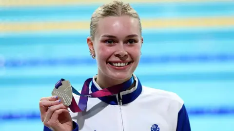Getty Images The swimmer Katie Shanahan holding her silver Commonwealth Games medal in front of the pool. She has blond hair, is grinning with delight and is wearing a white and blue athletic/tracksuit top.