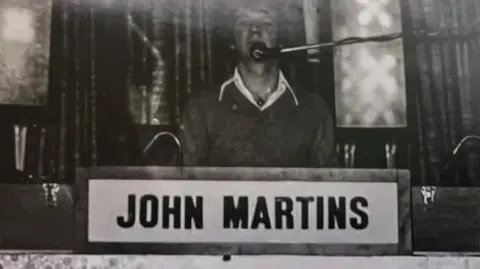 John Cripps A young John Cripps at a DJ booth in a black and white image. He has a microphone and the booth is labelled 'John Martins'.