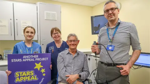 Spencer Mulholland Peter Russel and Kostas Marinakis are in a hospital room with two female nurses behind them. One of the nurses is holding a purple sign which says 'another stars Appeal Project' and has the Stars Appeal logo.
