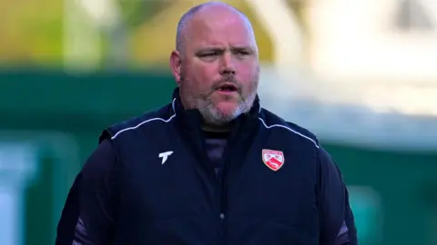 Outgoing Morecambe manager Jim Bentley watches on from the sidelines wearing a club-issued gilet 