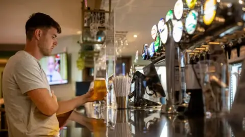 A side-on view of a bar in a pub. A barman is on the left, with brown hair and wearing a white t-shirt. He is holding a pint of beer