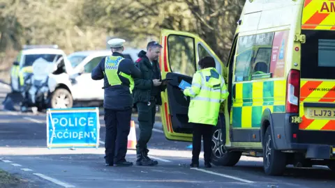 Paramedics and police gather with a crashed car in the distance