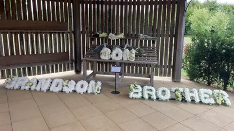Stephen Huntley/BBC Three flower wraiths are in the image. Two of them are on the floor with the one on the left saying grandson and the one on the right saying brother. There is another in the middle on a wooden bench which says son.
