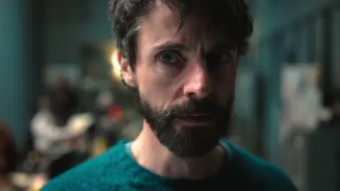 Close up of a man wearing a green jumper staring off to the right of the camera.