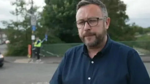 A mean wearing glasses and a dark blue shirt, with green railings and trees in the background as well as an individual wearing a high visibility vest