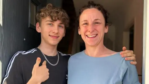 PA Media Jamie Holland standing in a doorway with his mother Carrie Holland. Both are smiling at the camera and Jamie is pointing to a silver chain around his neck.
