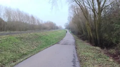 Cambridgeshire County Council A concrete path between two strips of grass. In the distance, two people are walking, one in a black jacket and another in a yellow one, but they are blurry.