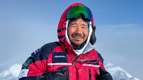 PA Media A man in a red snowsuit and a snow-coated beard smiles at the camera. There are snowy mountains in the backdrop. 