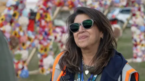 A woman with brown hair is wearing sunglasses and smiling. There are flowers behind her and she is wearing a high vis. 