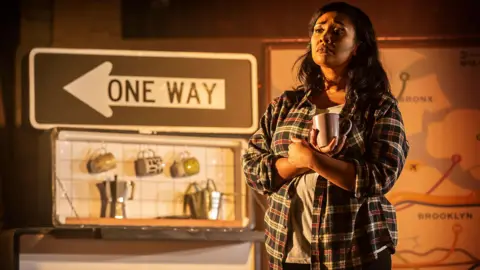 Pamela Raith Photography A woman, holding a coffee cup, wearing a plaid shirt and T-shirt, with long dark hair, looking out, with props that look like a small kitchen and a one way sign. The colours are muted. 