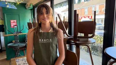 Becki Bowden / BBC Lydia has medium-length brown hair and a fringe. She is wearing a striped top and a dark green apron. She is standing next to the window and alongside the barricade. 