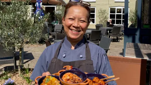 GB Hospitality Marketing A woman with brown hair tied back and glasses on her head. She is wearing a black chef's apron and is holding a tray of Thai food. She is outside and there is outdoor seating behind her, in front of a restaurant building