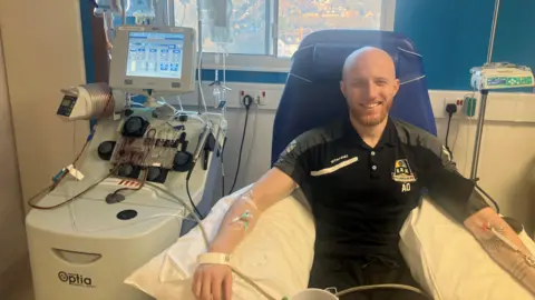 Bradford Teaching Hospitals NHS Foundation Trust Alex Overton sits in a chair in hospital connected to a series of drips, with medical equipment to the side of him. He is smiling. 