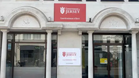 BBC The Government of Jersey building, one red sign in the centre of two pillared windows, in the between lower down a white sign saying the same