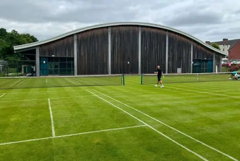 WLTSC Two outdoor grass tennis courts. A man wearing black shorts and a black t-shirt is walking towards a gap between the two courts, while holding a racket. A large wooden building with a curved silver roof is in the background.