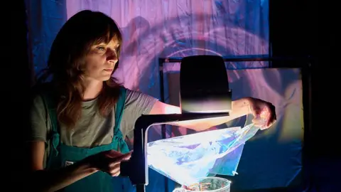 The Whiting's On The Wall Katie-anna Whiting drapes a plastic sheet on part of an overhead projector to create an image on a wall behind her. She has long brown hair and wears a T-shirt with dungarees. 