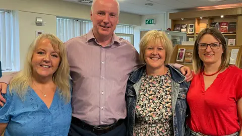 Left to right: Helen Roberts, who is wearing a blue embroidered short-sleeved blouse, and she has shoulder length blond hair, Nick Davies, who is the tallest in the group is wearing a stripey shirt and has short cropped grey hair, Margaret Spooner is stood next to him wearing a floral dress and a blue denim jacket. She has short blond hair and stood next to her is Faith Reynolds who is wearing a red v-neck t-shirt and blue jeans. She has short blond and brown balayage dyed hair and is wearing black glasses.  