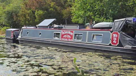 Lichfield & Hatherton Canals Restoration Trust A grey and red narrowboat is moored on a canal surrounded by trees, with lily pads and algae covering parts of the water's surface.