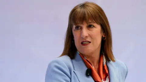 Reuters Rachel Reeves gritting her teeth with a lapel microphone attached to her pale blue jacket. She has shoulder-length brown hair and a fringe.