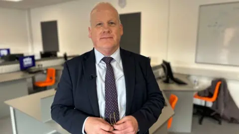 Kate Bradbrook/BBC Conor Renihan, standing in a classroom, with orange chairs in it, and a number of desks. He has short grey hair, is looking at the camera, holding a pair of glasses and is wearing a white shirt, with a tie, with spots on and is wearing a blue suit jacket. 
