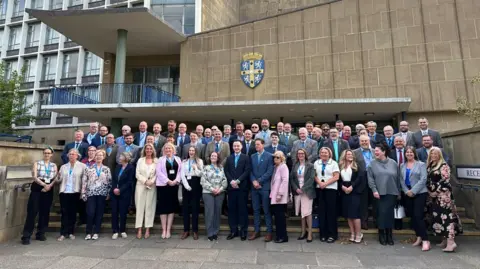 The new council members smiling as they line the steps in front of Durham County Hall in May 2025. It is a beige building with a lot of windows and the county crest.

