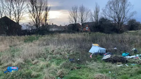 BBC/Stuart Whincup A overgrown grassy area with fly-tipped waste. There are trees and houses in the distance.