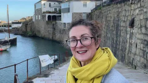 A woman smiles at the camera she wears glasses and a bright yellow scarf.
Behind her is a slipway leading into Newlyn Harbour where boats are moored up.
