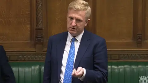 UK Parliament Oliver Dowden speaking in Parliament, wearing a blue speckled tie and blue suit with white shirt. He has the green leather parliamentary bench behind him and is gesturing with his hand to make a point. 