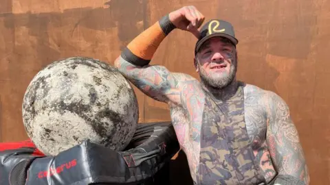 Shaun Slicker with elbow resting on big boulder. He has tattoos and a combat style vest.