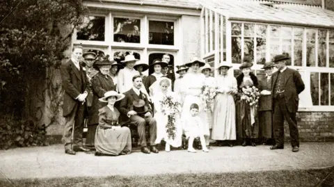 Hestercombe House Trust A black and white image showing a group of around 20 adults, standing up, in old-fashioned formal clothes, including wide-brimmed hats, long dresses and suits. There is a couple sitting in front of them, with a man holding a top hat in his lap and a woman in a wedding dress. She and several of the standing women are holding large floral bouquets. Behind the group is an orangery with many glass panes. 