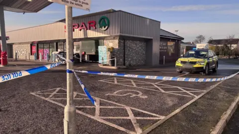 A police cordon at Spar filling station - there is blue and white police tape in the foreground wrapped around a lamppost and a police vehicle is just behind it. Beside it is the large Spar shop with grey cladding and a Spar logo.