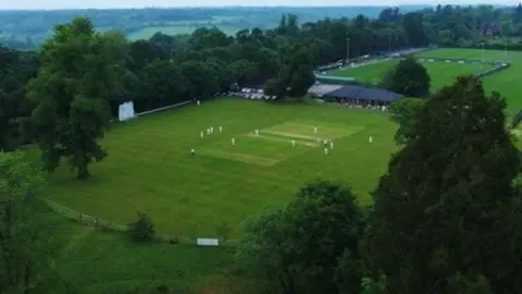 Reigate and Banstead Borough Council An aerial view shows the ground at Chipstead, Coulsdon & Walcountians Cricket Club with players on the pitch, surrounded by green fields, trees and a pavilion building.