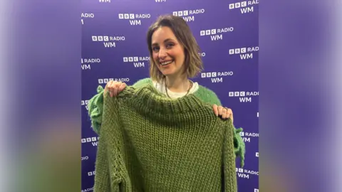 A woman with short brown hair grins at the camera as she holds up a green knitted jumper. She stands in front of a purple BBC Radio WM background.
