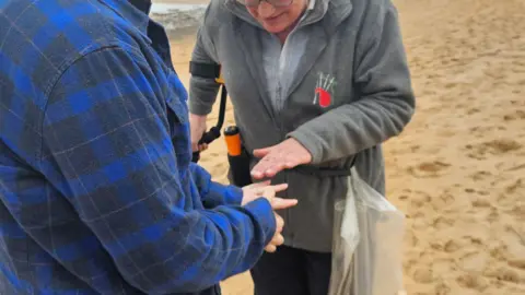 A woman in a blue checked top showing a woman in a grey jumper her engagement ring. 