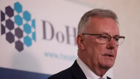 PA Media Health Minister Mike Nesbitt speaking to media at the Department of Health, Castle Buildings, Stormont Estate, Belfast. He has short grey hair, wearing glasses, a dark blazer and white shirt. Department of Health branding is behind him.