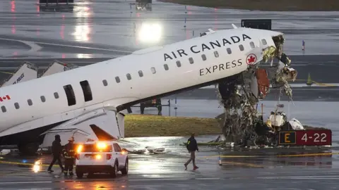 Air Canada flight AC8646 with its nose upturned and wrecked. Emergency services surround it. The tarmac is wet, it is early morning.