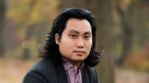 Es Lee con un blazer gris y una camisa a cuadros Es Lee se ve en la cámara.