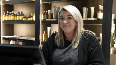 Photograph of Katie Bishop, aged 30. She stands behind the till inside Goji hair salon. She has bold below shoulder length hair. She has brown eyes and smiles at the camera. Shelves can be seen in the background, which are down lit with lights and have lots of products. 