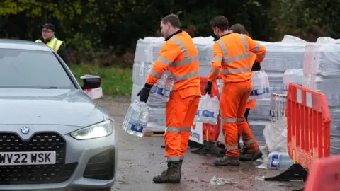 PA Media Several people wearing hi-vis orange jackets are lifting slabs of water bottles at a car park.