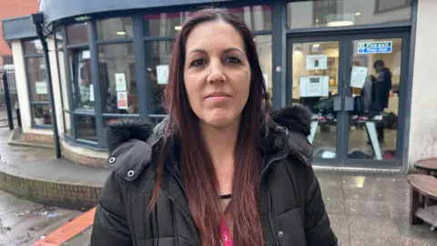 Joanne Pritchard is standing outside the Hope Centre’s day facility on a wet pavement, with the curved glass frontage of the building visible behind them. The entrance doors, noticeboards and interior lights can be seen through the windows, where people are gathered inside the warm space. The person is dressed in a winter coat suitable for the cold, rainy weather, and the surroundings include a small seating area and the brickwork of nearby buildings, reflecting the busy, urban setting of the support hub.