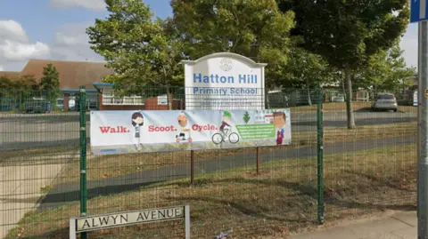 Google A view of the grounds of a primary school surrounded by a green wire fence. Immediately behind the fence is a white signboard with the words 'Hatton Hill Primary School' in blue lettering.