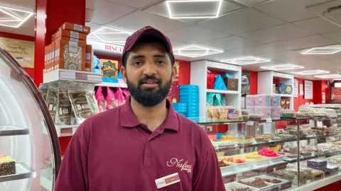 Faizal Mahmood wearing a maroon polo shirt with the name 'Nafees' embroidered and a maroon cap. Behind him are several shelves of cakes and desserts behind glass as well as shelves with boxes of biscuits and cakes. 